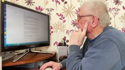 An elderly gray-haired man with glasses sits at a table in front of a computer Stock Footage 233570473