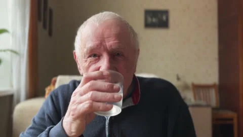 An elderly gray-haired man with a happy face drinks water from a glass at home Stock Footage 233570755