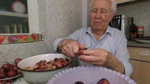 An elderly gray-haired man sits at the kitchen table and cuts fruit. Stock Footage 137787218