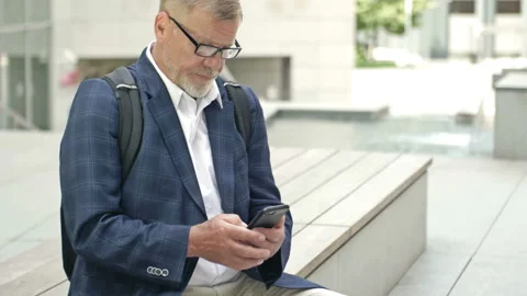 Elderly grey-haired man using smartphone while resting on street, sitting on Stock Footage 159272829