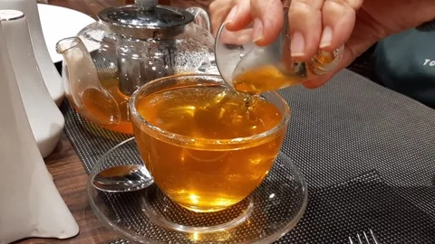 Elderly Hand Pouring Thick Liquid into Herbal Tea with Honey on Wooden Table Stock Footage 317945373