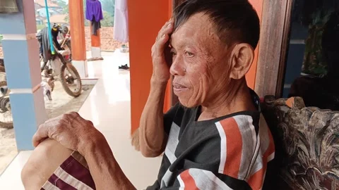 An elderly Indonesian man sits on his porch with his hand on his forehead, Stock Footage 315308853