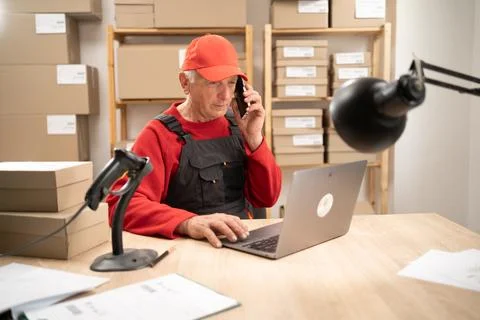 Elderly logistics worker using laptop and smartphone indoors Stock Photos