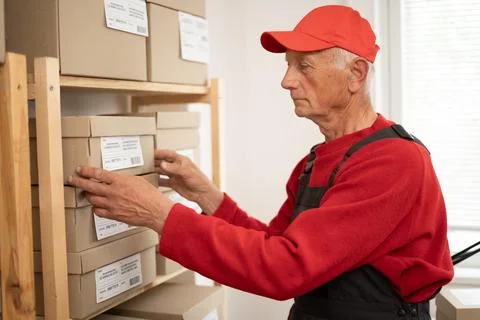 Elderly mail worker applying delivery address label on parcel box inside po.. Stock Photos