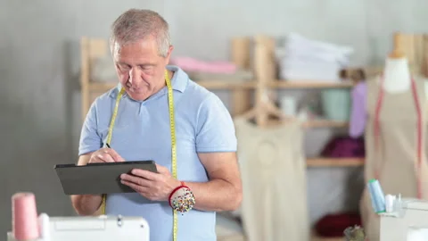 Elderly male tailor draws pattern using tablet in sewing workshop Stock Footage 315032815