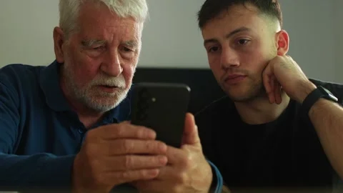 An Elderly Man and Young Man Intently Look at a Smartphone Screen, Sharing a Mom Stock Footage 335240564