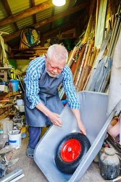 An elderly man with beard in work clothes assembles a wheelbarrow in a workshop. Stock Photos