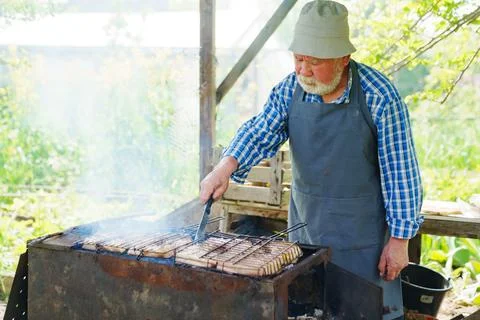 An elderly man with a beard in work clothes cooks meat steaks on the grill Stock Photos