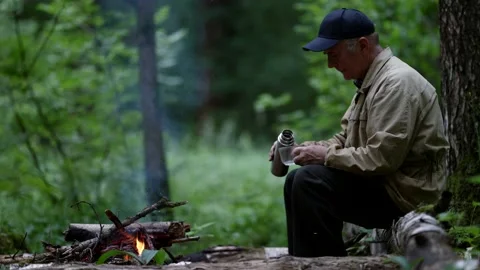 An elderly man in a cap is sitting in the forest on a log in front of a fire and Stock Footage 163664911