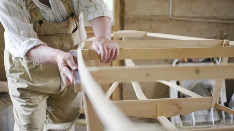 Elderly man carpenter builds a small boat with his hands out of wood in a small Vídeos de archivo 89787221