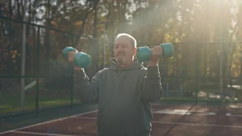 An elderly man doing exercises with dumbbells on a sports field. Fitness for Stock Footage 255295085