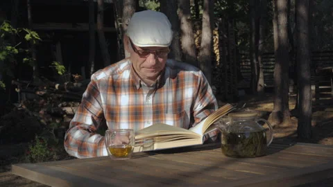 Elderly man enjoying tea while reading a book in a peaceful wooded setting Stock Footage 289297546