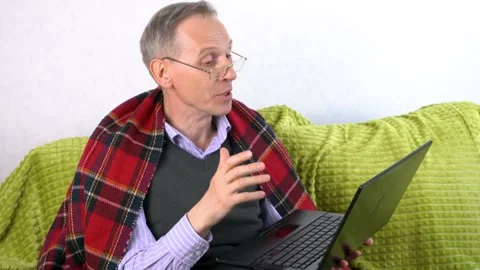 An elderly man with glasses, covered with a red blanket, talks on Skype, sitting Stock Footage 300377747