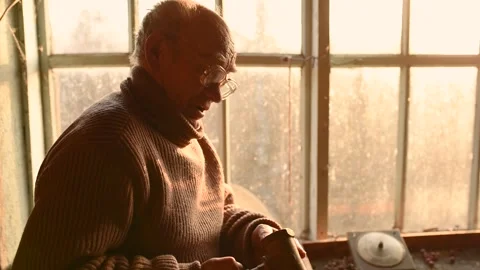 Elderly man glasses picks up old stuffs brushing dust off in attic house window Stock Footage 190767734