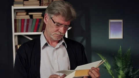 An elderly man is glasses is standing in front of the bookshelf with a book in Video stock 132635937