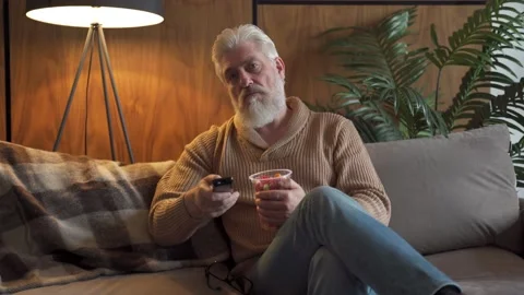 An elderly man with a gray beard sitting on the sofa switches channels on the TV Video stock 142753564