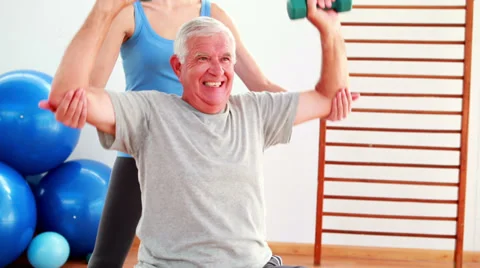 Elderly man lifting hand weights sitting on exercise ball with his trainer Video stock 34130554