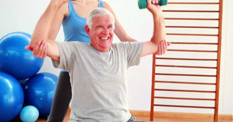Elderly man lifting hand weights sitting on exercise ball with his trainer Video stock 34248202