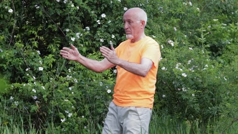 An elderly man performs knee strikes on his palms against a natural backdrop Stock Footage 310890577