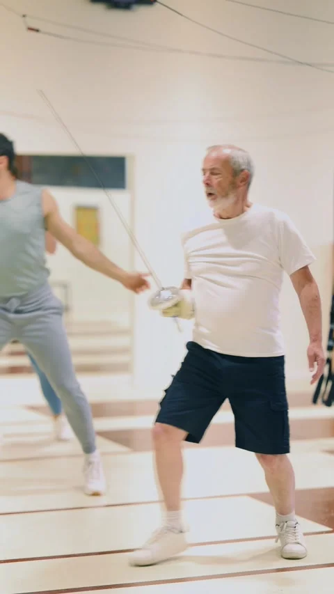 Elderly man practices fencing techniques with a group of other athletes in a gym Stock Footage 346119406