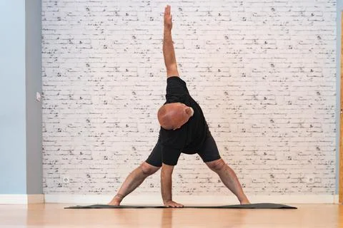 Elderly man practicing a triangle pose yoga on a mat in a bright, spacious room. Fotos Stock