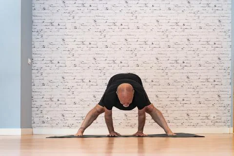 Elderly man practicing a wide-legged forward bend yoga pose on a mat in a bright Stock Photos