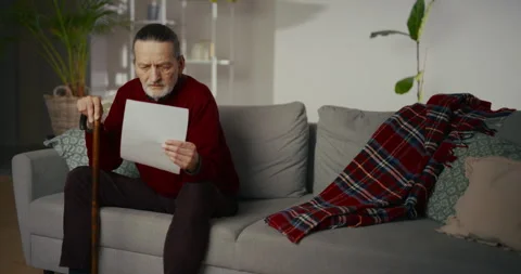 Elderly man reading a document while sitting on a sofa in a living room Vídeos de archivo 336286759