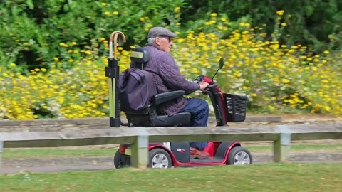 Elderly man riding electric mobility scooter through a park in England, UK. Stock Footage 202128624