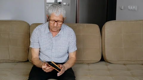 An elderly man sits on a couch and examines old photos. Good memories. Stockbeeldmateriaal 111063994
