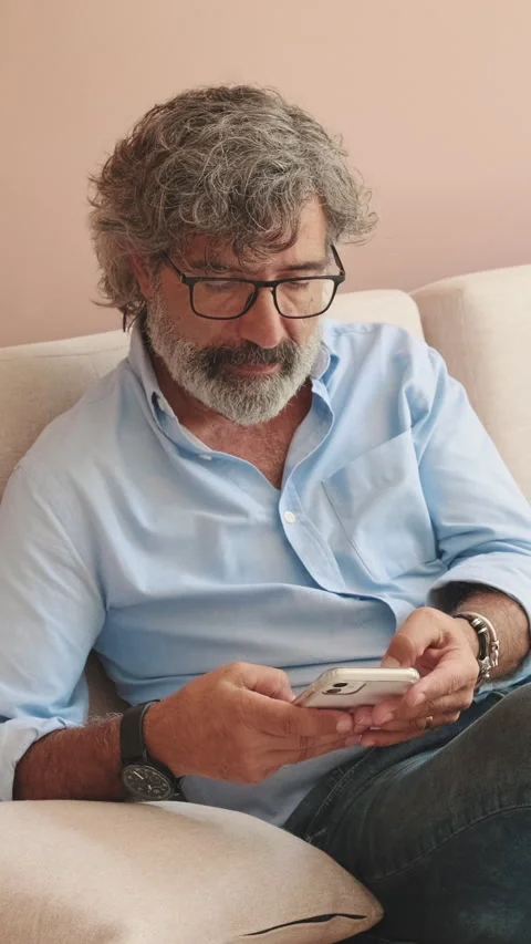 An elderly man sits on sofa in cozy living-room with phone in his hands Stock Footage 252502427