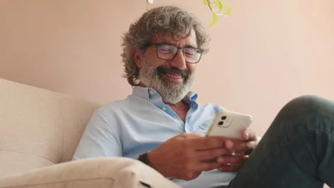 An elderly man sits on sofa in cozy living-room with phone in his hands Stock Footage 253928382