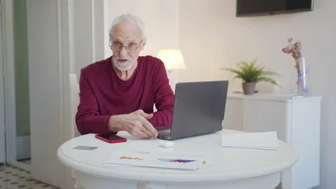 An elderly man sits at a table with a laptop and says serious and important Stock Footage 173958929