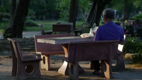 Elderly man sitting alone at a red concrete park table Stock Footage 332293300