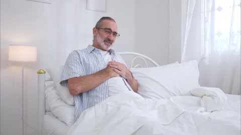 Elderly man sitting on the bed and holding his chest in pain 스톡 동영상 326179008