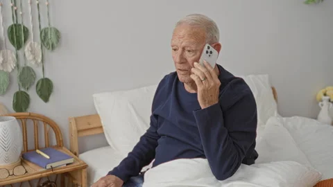 Elderly man sitting on bed holding smartphone to ear, hands raised in exasp.. Stock Footage 337377030