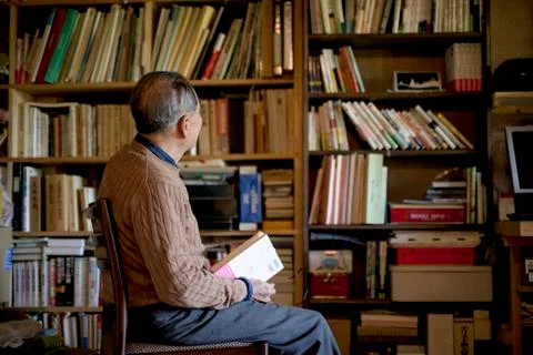Elderly man sitting on chair in front of bookcase, holding book. Stock Photos