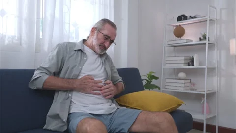 An elderly man sitting on a sofa holding his stomach in pain 스톡 동영상 326179808