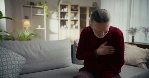 Elderly man sitting on sofa holding chest in bright living room Видео 336287045