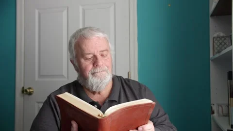 An elderly man is sitting at a table and reading a book. Sunlight comes through Stock Footage 338057903