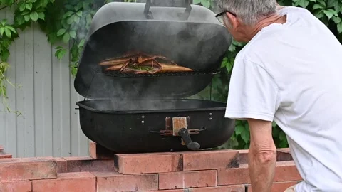 An elderly man smokes fish on a brick barbecue Stock Footage 134341892