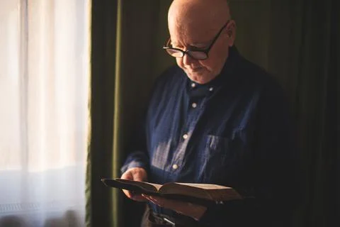 Elderly man stand next to window and reading Bible Stock Photos