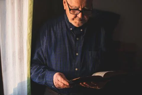 Elderly man stand next to window and reading Bible Stock Photos