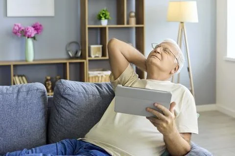 Elderly man with tablet on couch at home, stretching neck and relaxing indoors Stock Photos