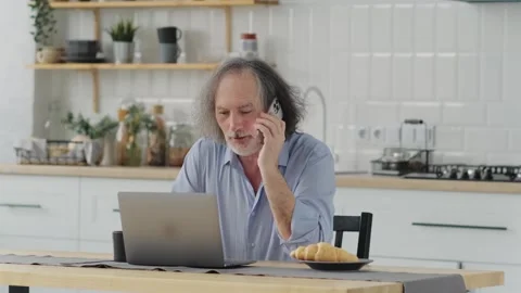 An elderly man talking on the phone while sitting at home near a laptop, smiling Stock Footage 243975647