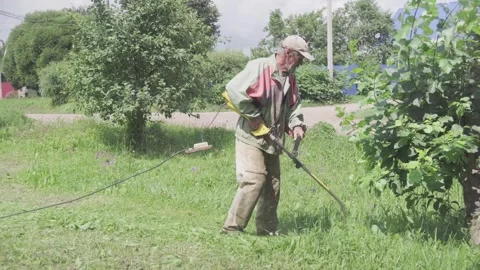 Elderly man trims grass with electric trimmer in his yard on a sunny day Stock Footage 330397706