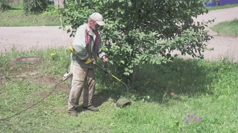 Elderly man uses electric trimmer to cut grass in yard on a sunny day Stock Footage 330397751