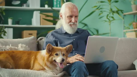 Elderly Man Uses Laptop Computer, While Sitting on a Couch with His Lovely Corgi Stock Footage 196243161
