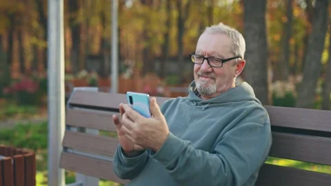 An elderly man uses a smartphone while sitting on a park bench. Pensioner Stock Footage 255714903
