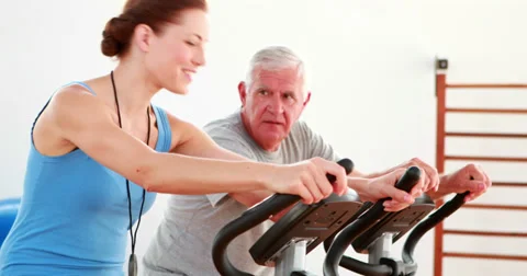 Elderly man using the exercise bike with his trainer Stock Footage 34248613