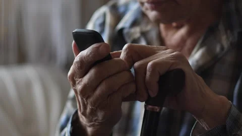 An elderly man with a walking stick uses a push-button telephone while sitt.. Vídeos de archivo 288350448